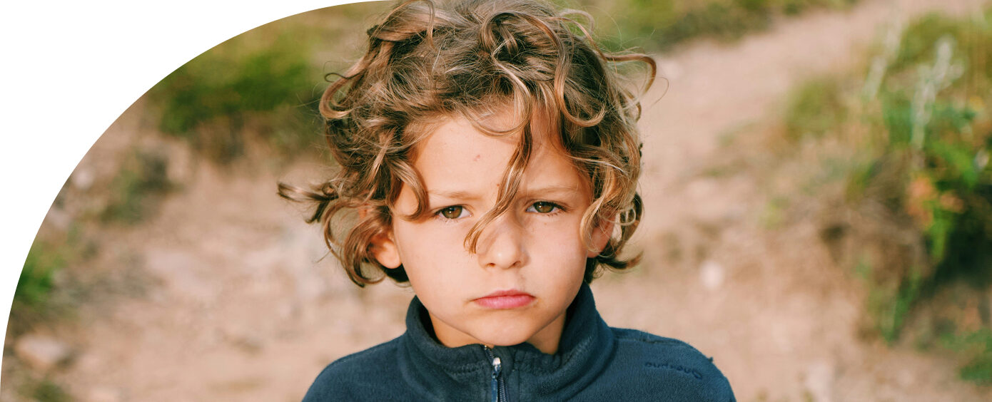 Young child with curly hair and serious expression, wearing a blue jacket, standing outdoors on a dirt path.