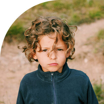 Child with curly hair in a blue jacket standing outdoors, looking serious.